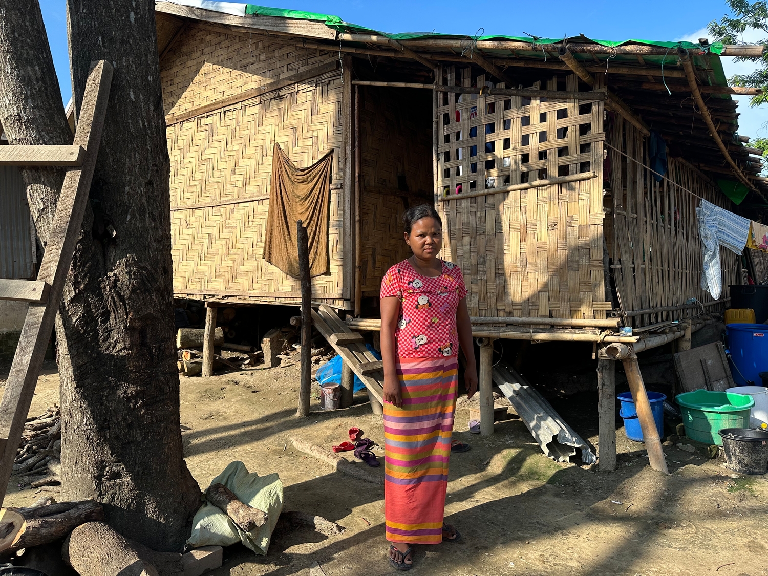 A woman stands in front of a bamboo shelter on stilts.